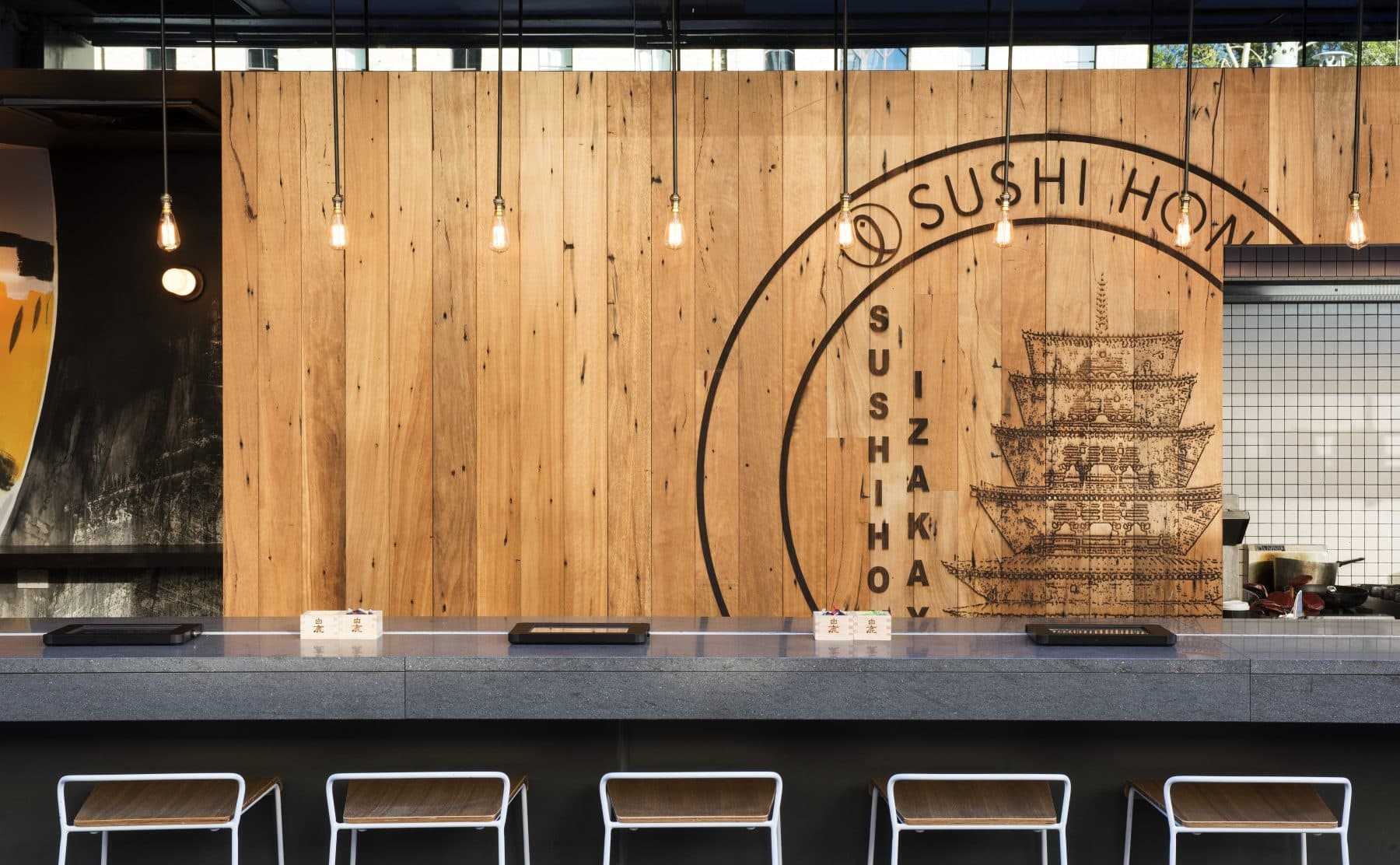 An empty sushi restaurant counter with wooden wall, Edison bulb lighting, and a SUSHI HOZ ship-illustration sign