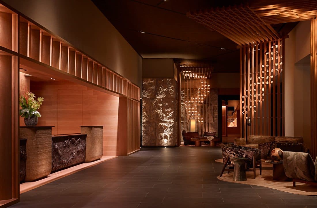 A modern Japanese hotel lobby with a wooden reception desk, stone wall features, and a warm lounge area with hanging lights