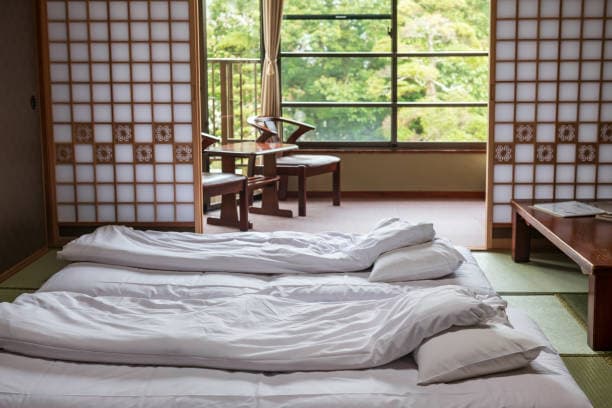 A traditional Japanese ryokan guest room with futon bedding on tatami mats, low wooden table, shoji screens, and a garden view